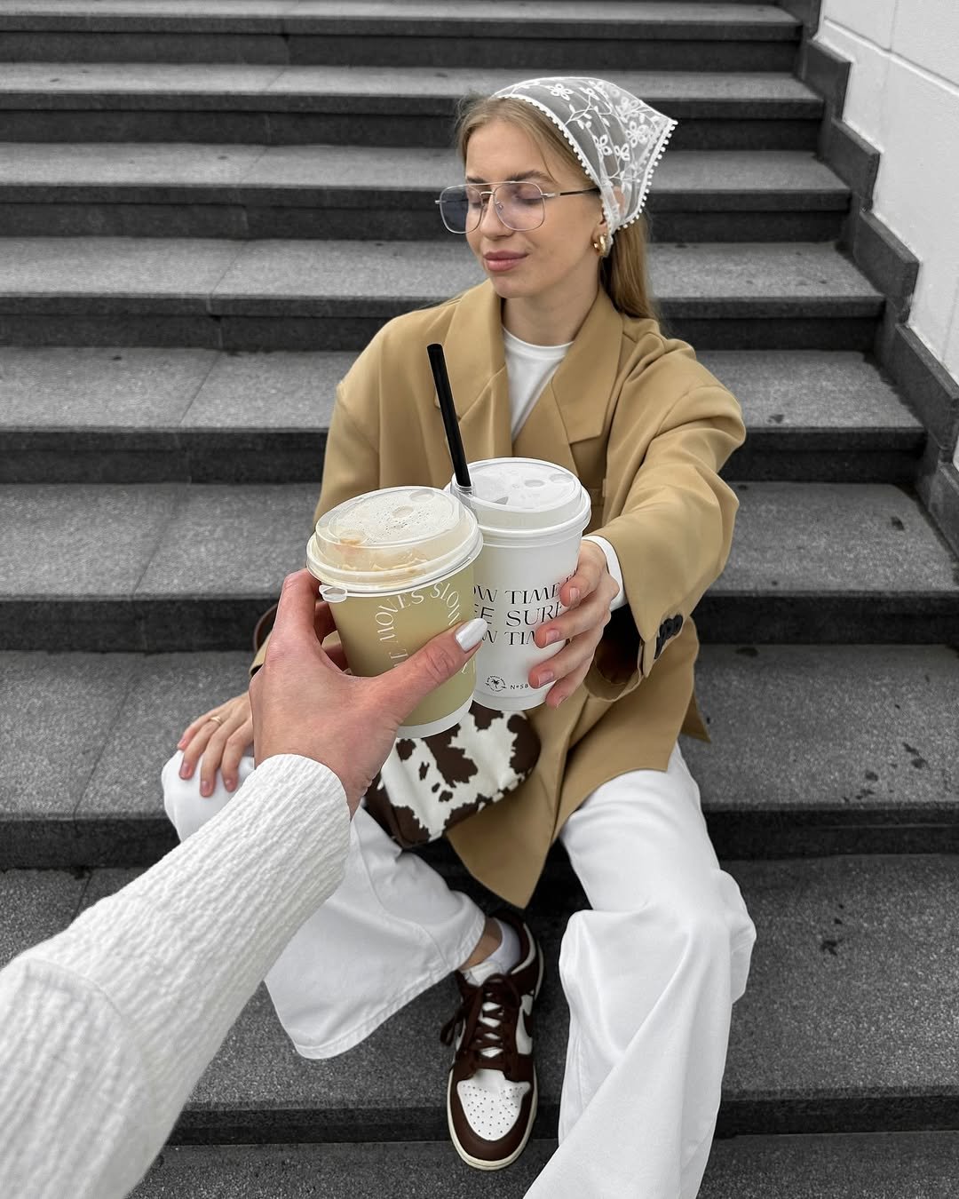 A jovem está sentada numa escada, segurando um copo de bebida e usando um lenço de renda na cabeça.