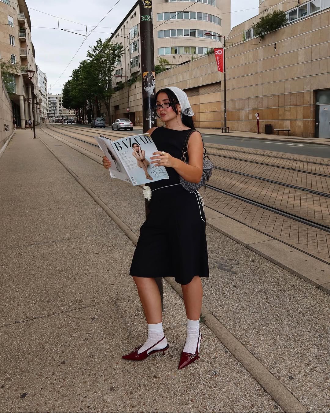 Pessoa lendo revista em calçada urbana, usando vestido preto, sapatos vermelhos e lenço de renda na cabeça.