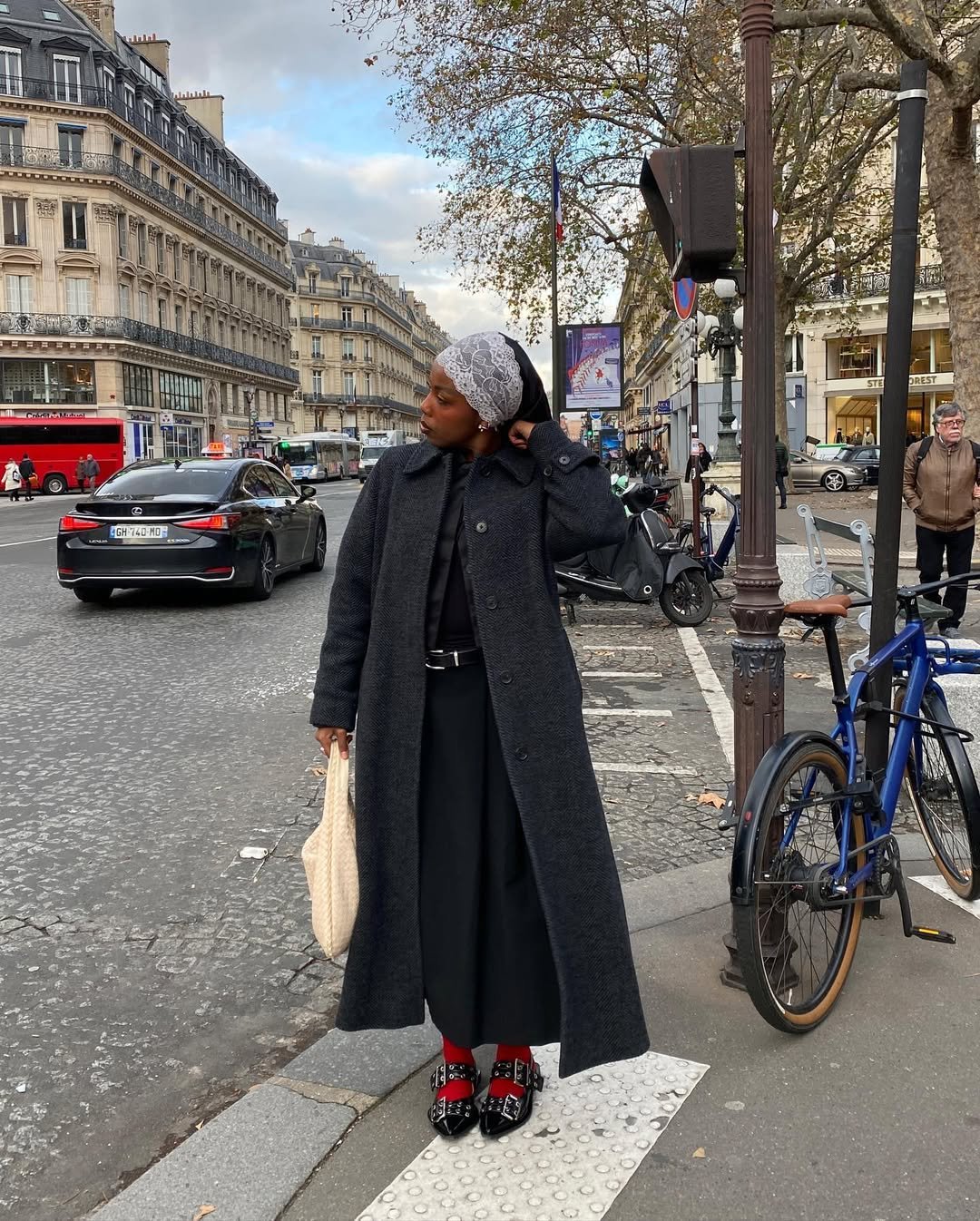 Pessoa com lenço de renda na cabeça usa casaco escuro em rua movimentada de cidade europeia.
