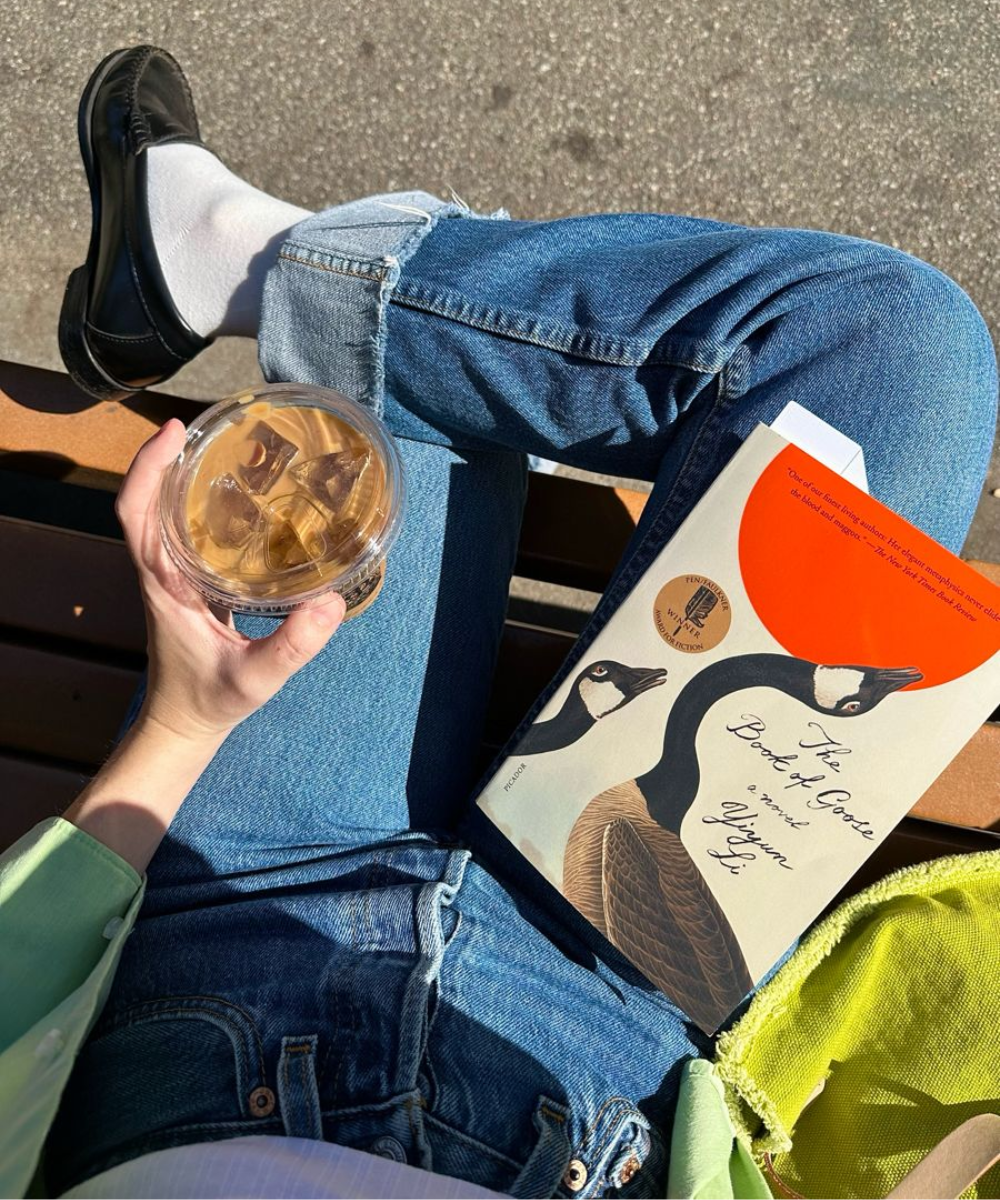 Pessoa sentada ao sol com jeans e mocassins, segurando bebida gelada ao lado de livro. Livros que as celebridades amam.