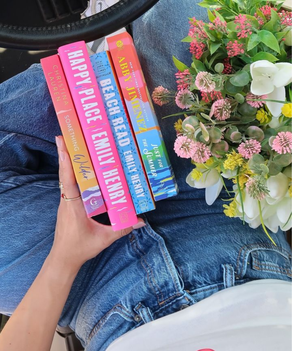 Pessoas segurando livros coloridos sobre romance e fantasia ao lado de flores vibrantes no colo.