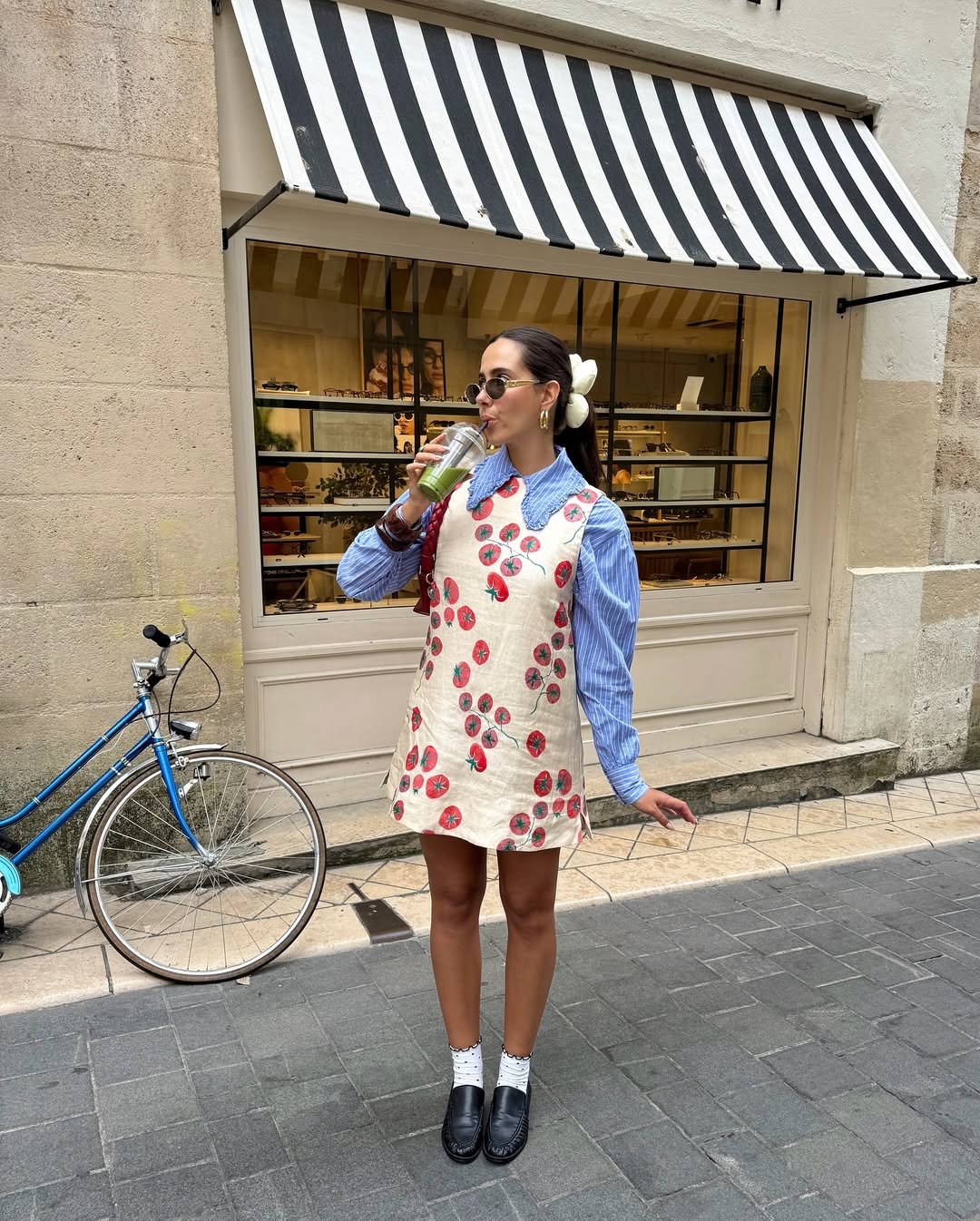Pessoa toma suco verde e usa vestido estampado, camisa listrada azul e loafer feminino preto em rua pavimentada.