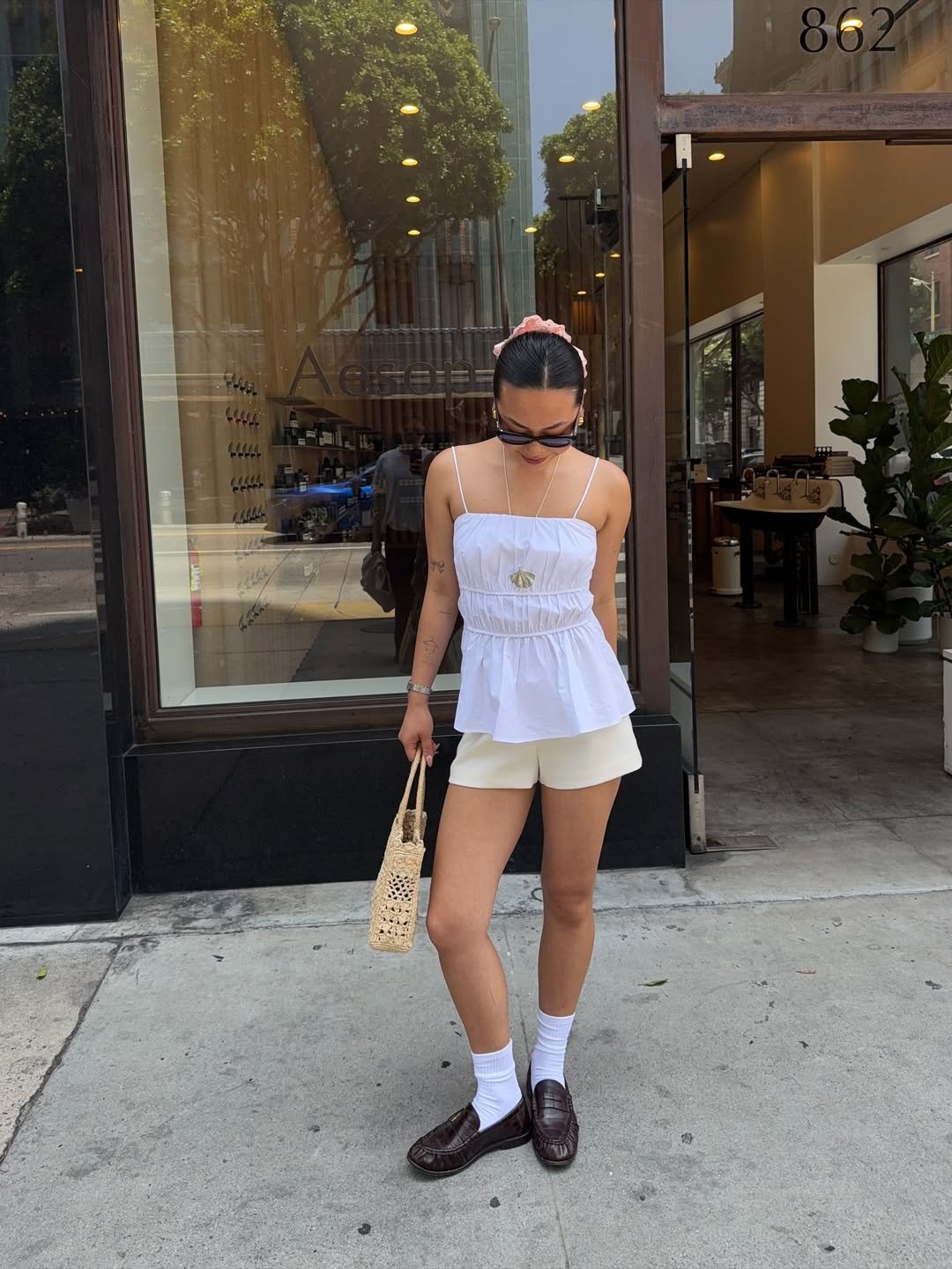 Pessoa com blusa branca e shorts bege, usando loafers femininos escuros, bolsa de palha e óculos escuros.