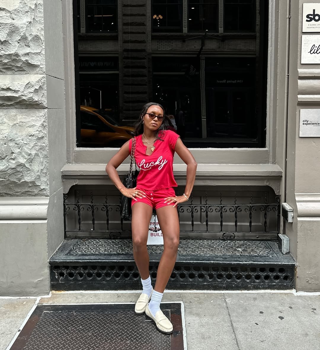 Pessoa de óculos usa roupa vermelha e loafer feminino branco, em frente a um prédio com escadas metálicas.