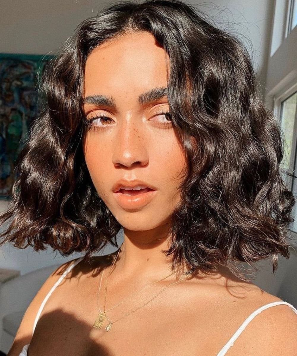 Mulher com cabelo ondulado escuro e curto, iluminada por luz natural, usando colar dourado.