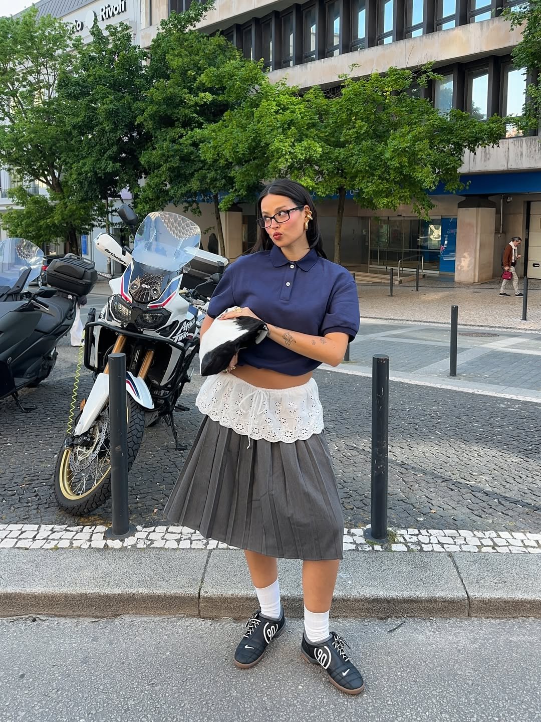 Mulher com camiseta azul e saia cinza em frente a uma moto, edifício e árvores ao fundo.