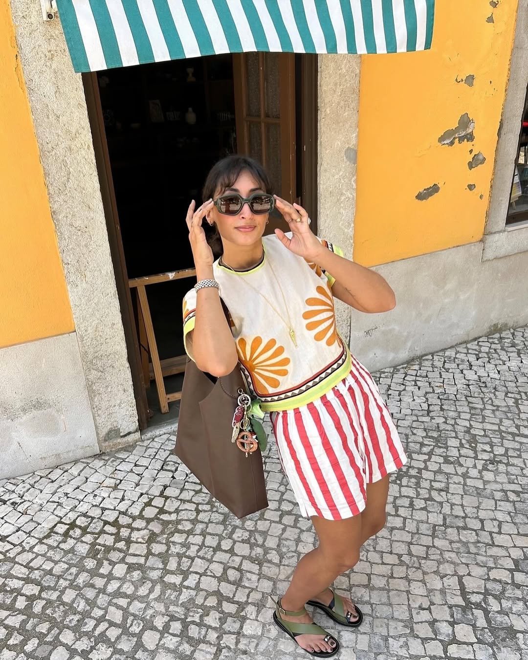 Pessoa com roupa colorida listrada e sandálias, óculos de sol em frente a uma parede amarela. Looks de calor estilosos.