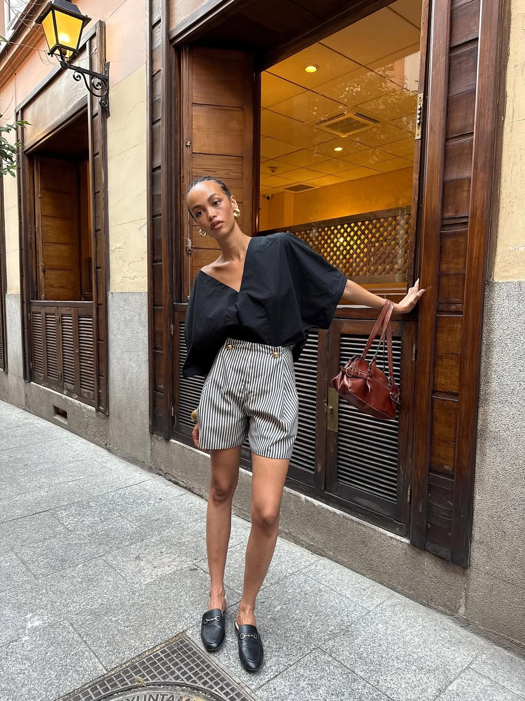 Pessoa em frente a um café, vestindo blusa preta larga e shorts listrados. Looks de calor estilosos.