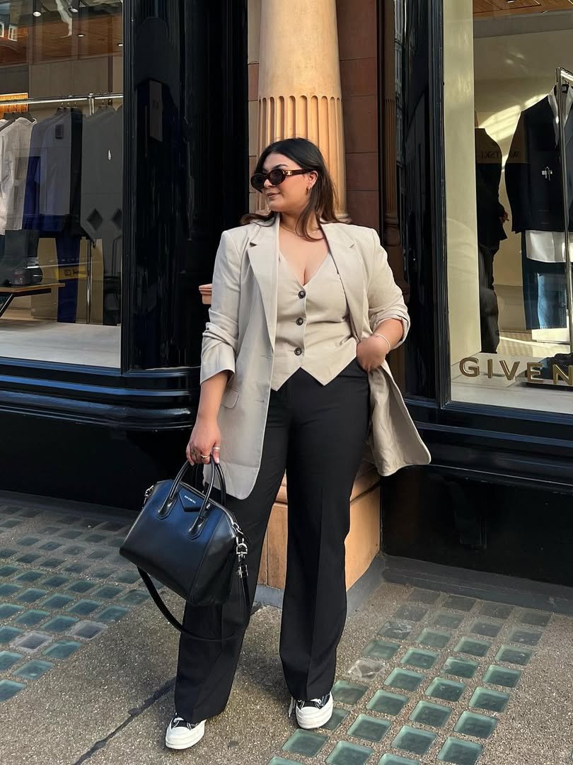 Pessoa com traje elegante, blazer e all star preto, em frente a loja. Carrega bolsa preta e usa óculos escuros.