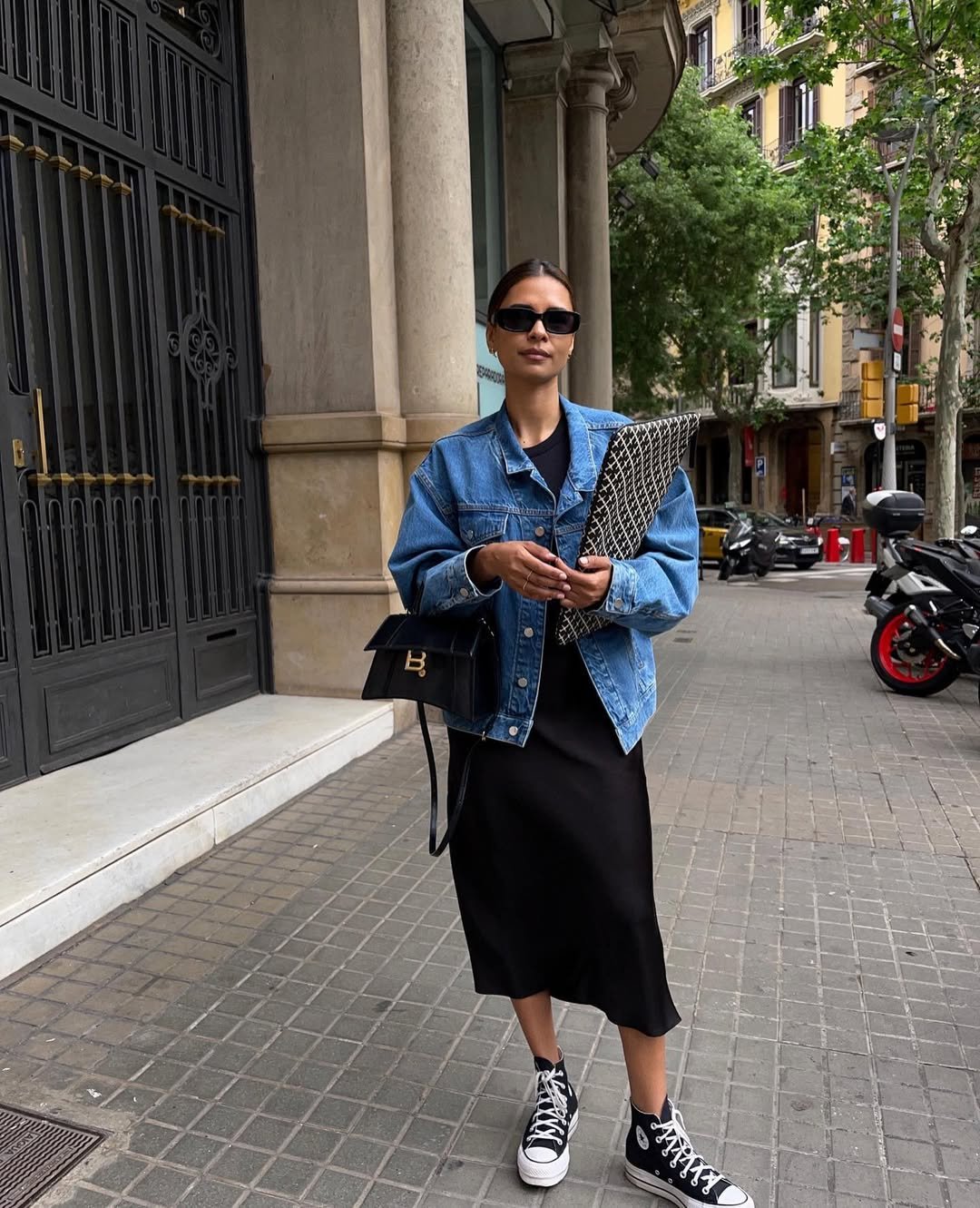 Pessoa usando jaqueta jeans, saia preta e tênis all star preto caminha na calçada de uma cidade com prédios e árvores ao fundo.