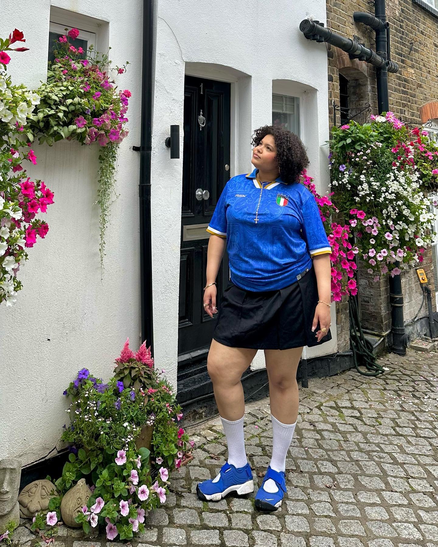 Pessoa vestindo camisa de time azul e saia preta, cercada por flores coloridas em uma calçada de paralelepípedo.