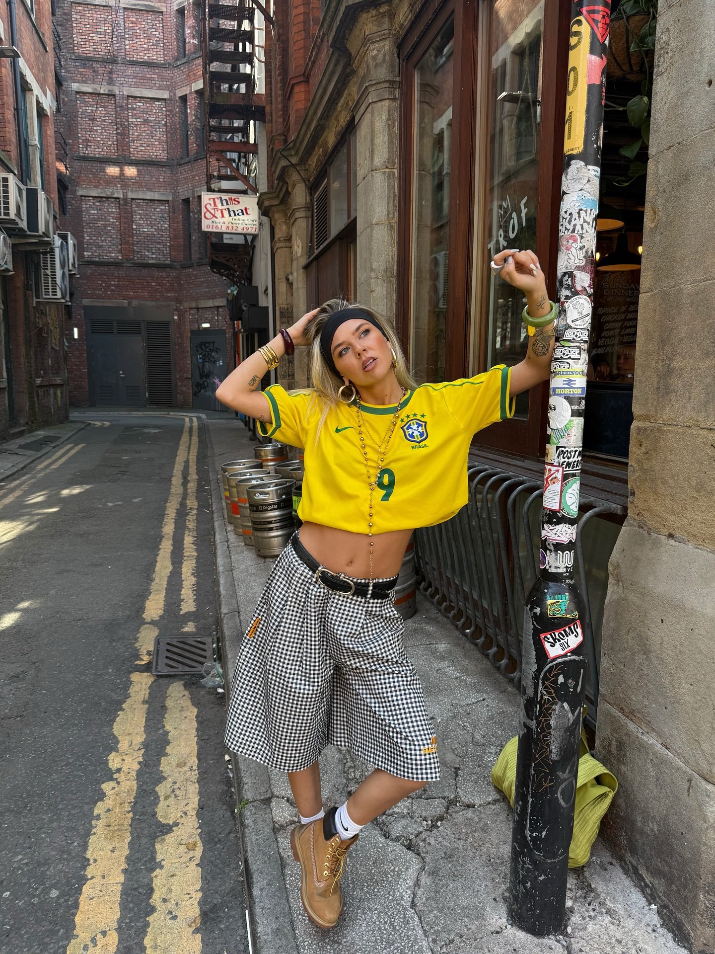 Pessoa usando camisa amarela de futebol e bermuda xadrez em uma rua de paralelepípedo. Representando looks com camisa de time.