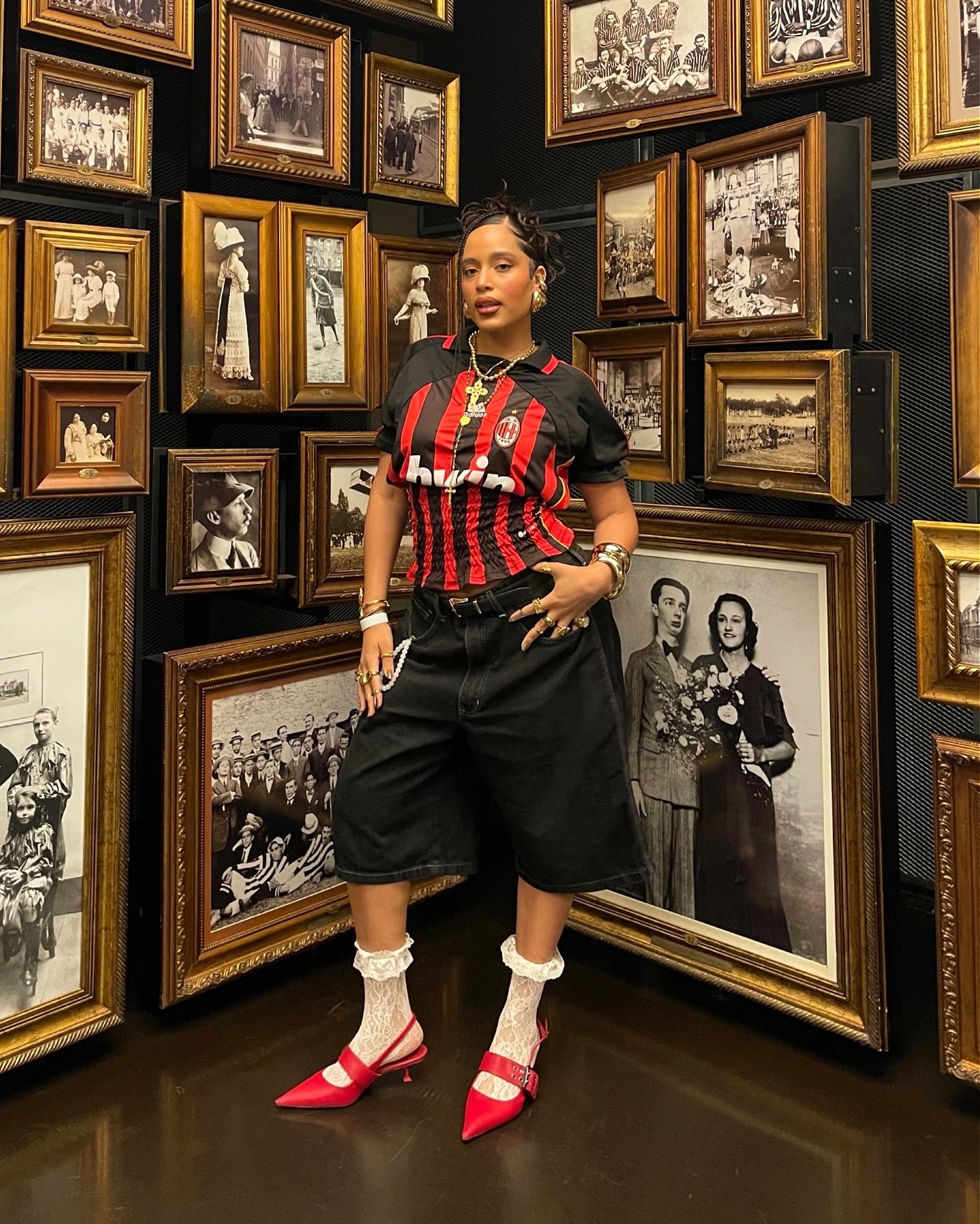 Mulher usando camisa de time listrada preta e vermelha, bermuda preta e sapatos vermelhos, em sala com quadros antigos.