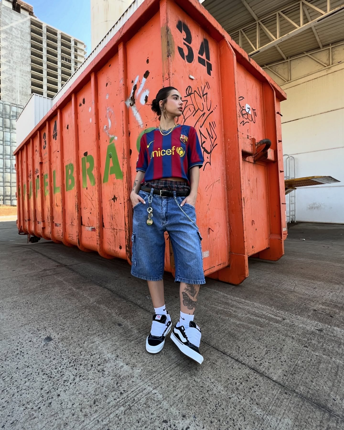 Jovem com camisa de time, bermuda jeans e tênis, ao lado de um grande contêiner laranja com grafites.