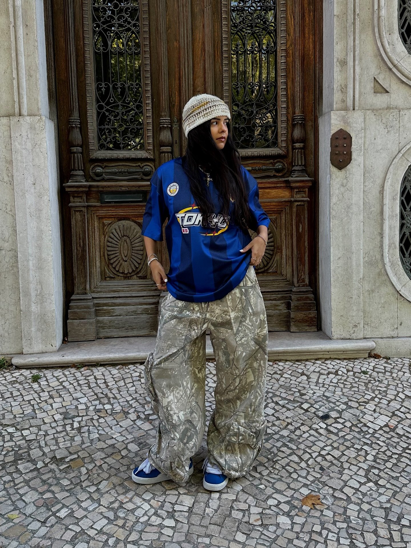 Pessoa usando camisa de time azul e calça camuflada, em frente a porta ornamentada.
