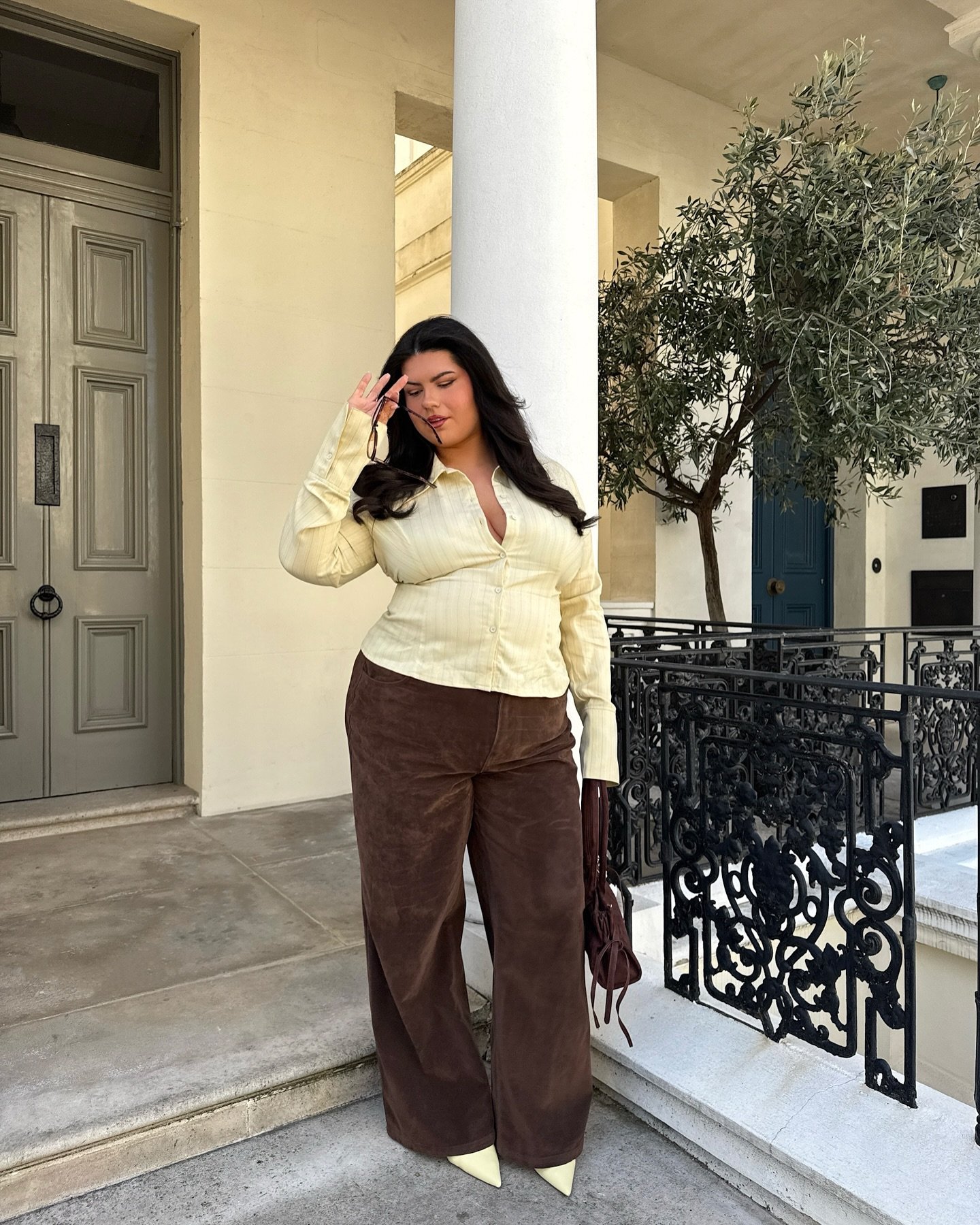 Usuária com camisa amarela e calça marrom em varanda com planta. Usa flats no look casual.