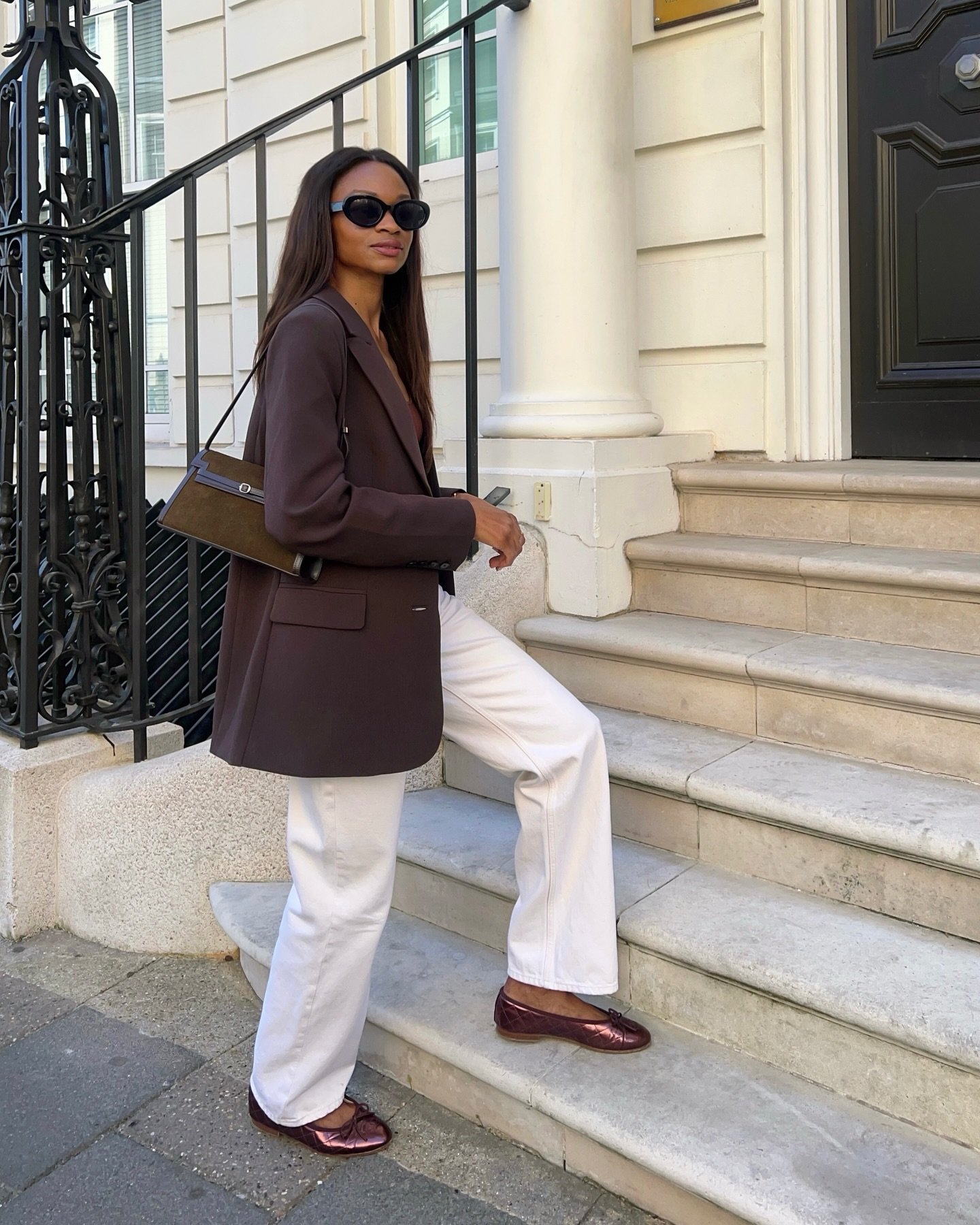 Mulher em casaco marrom e calça branca, usando sapatilhas metálicas e óculos escuros, sobe escadas ao ar livre.