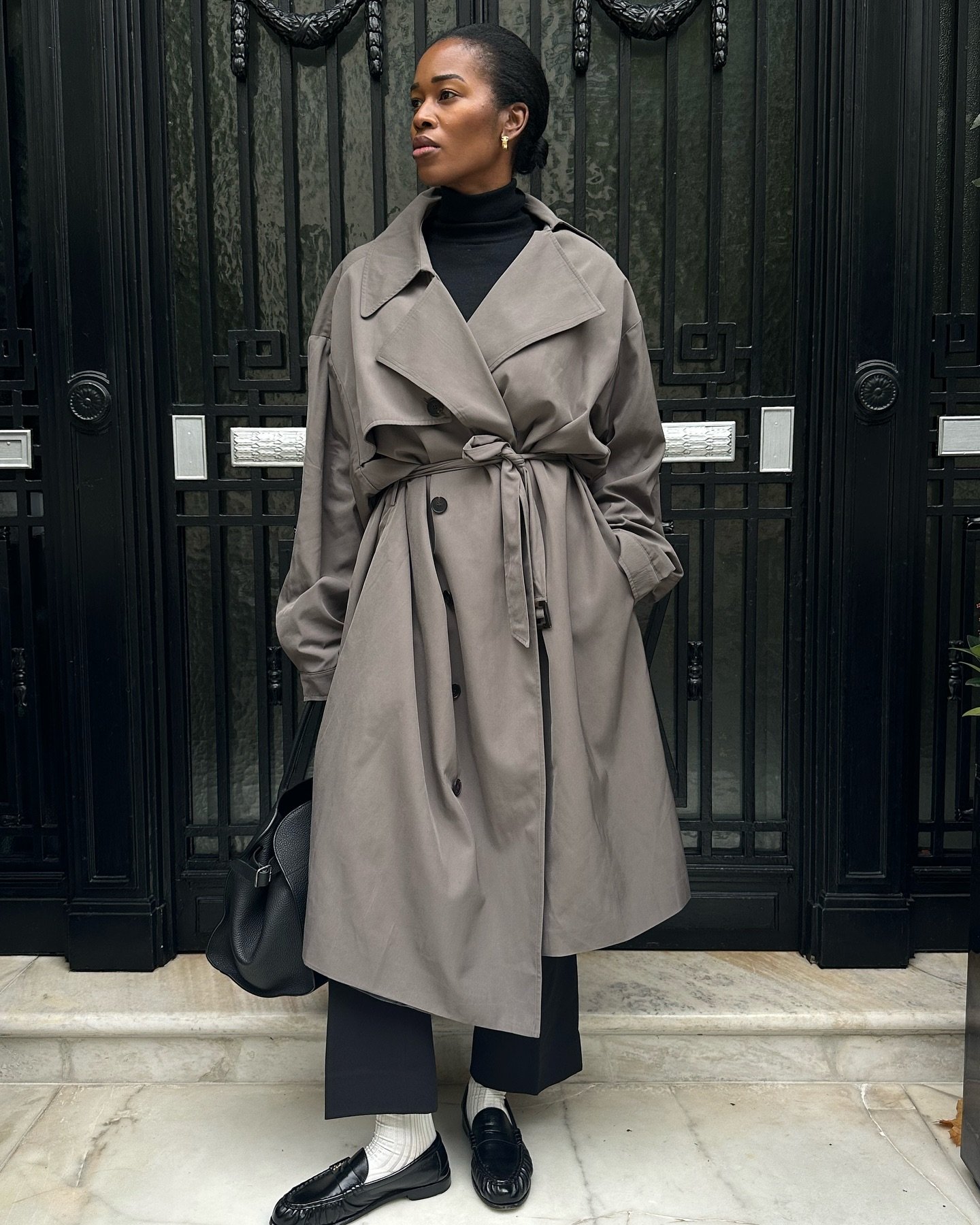 Pessoa com casaco cinza, calça escura e mocassins pretos, segura uma bolsa em frente a portas pretas e decorativas.