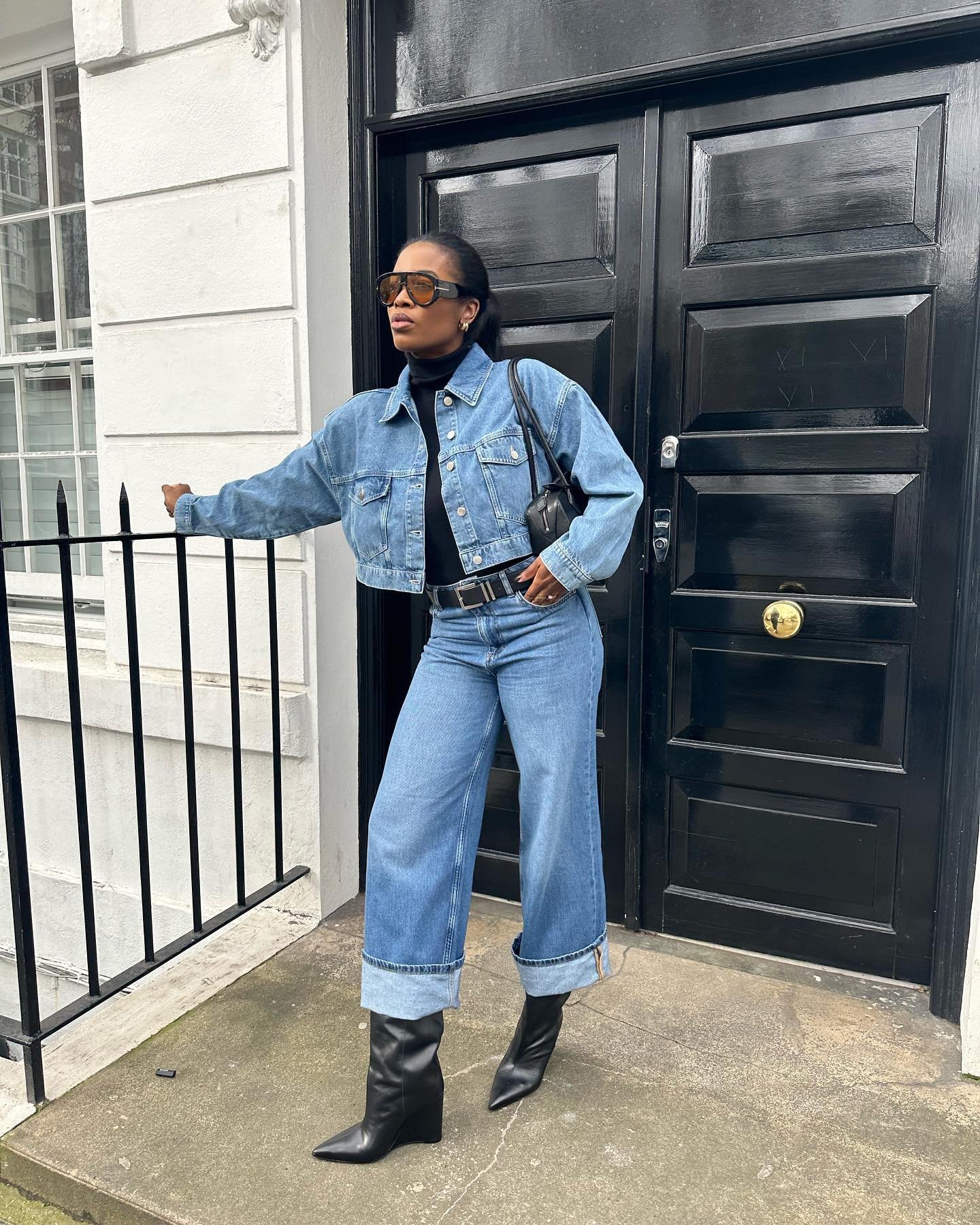 Vestindo jaqueta e calça jeans, pessoa usa óculos escuros e botas com salto anabela em frente à porta preta.