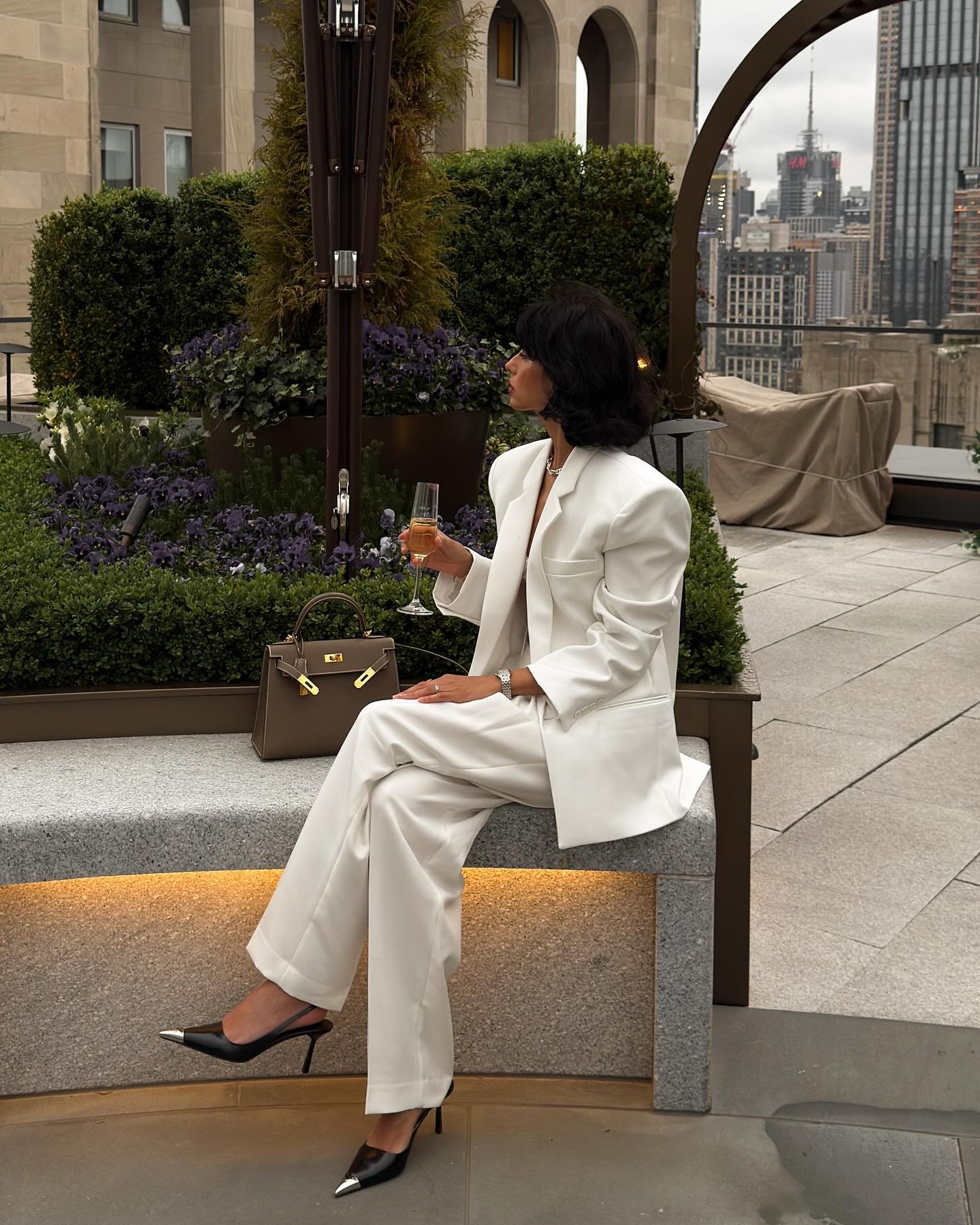 Mulher de terno branco elegante, segurando uma taça de champanhe em um terraço urbano sofisticado.