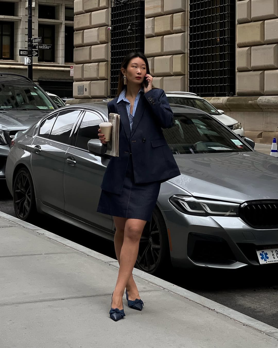 Mulher com look elegante formal, segura café e jornal, usando blazer e sapatos, em frente a carro estacionado.