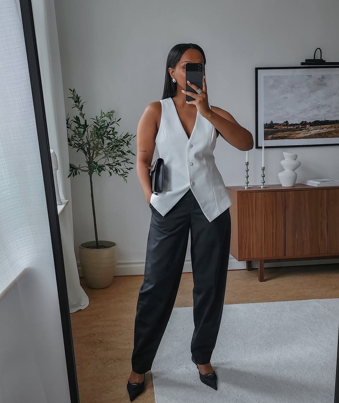 Pessoa em look elegante com colete branco e calça preta, segurando uma bolsa, em frente a uma cômoda moderna.