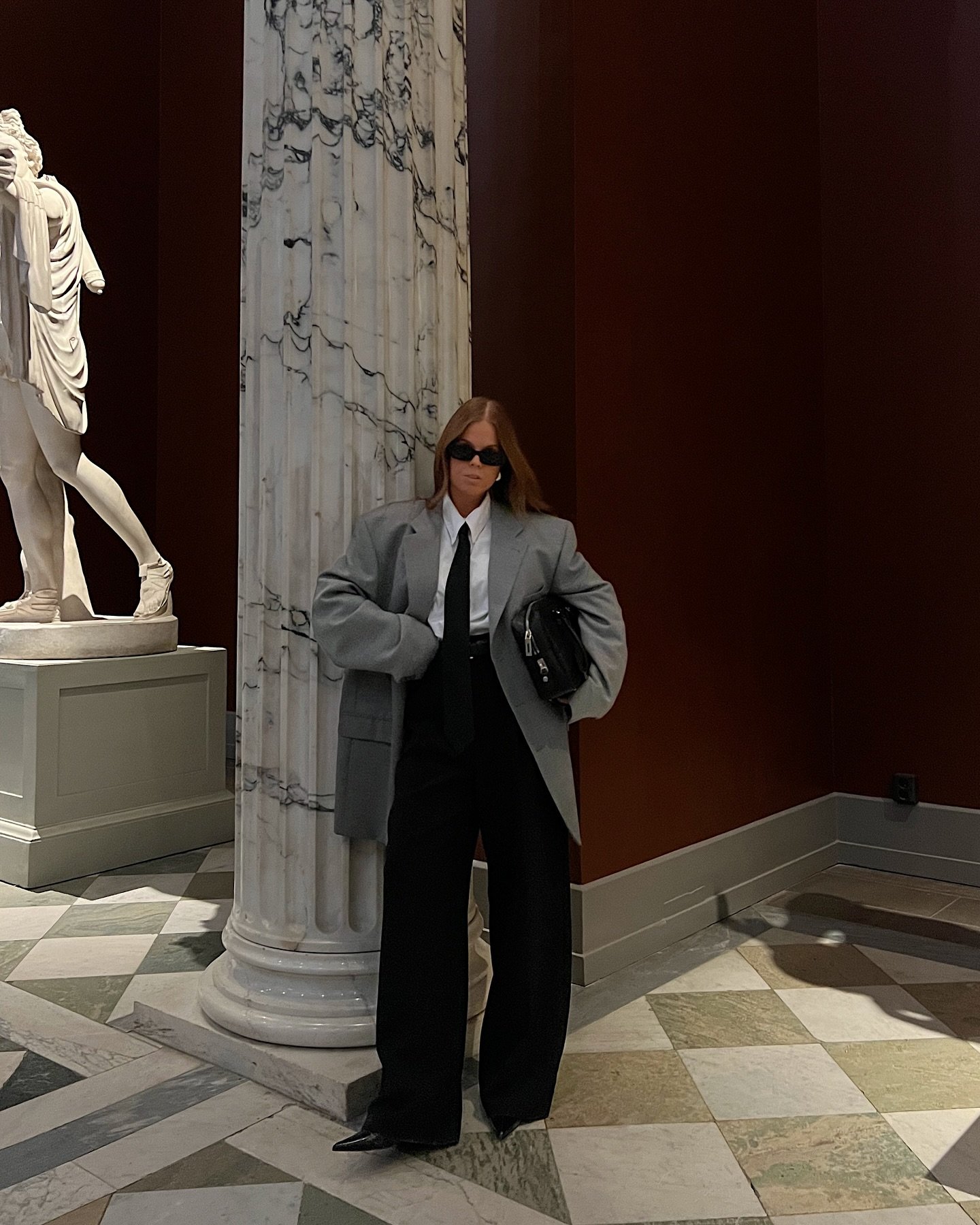 Pessoa em traje formal, óculos escuros e casaco cinza, em ambiente clássico com colunas e estátua. Representando looks elegantes para trabalhar.