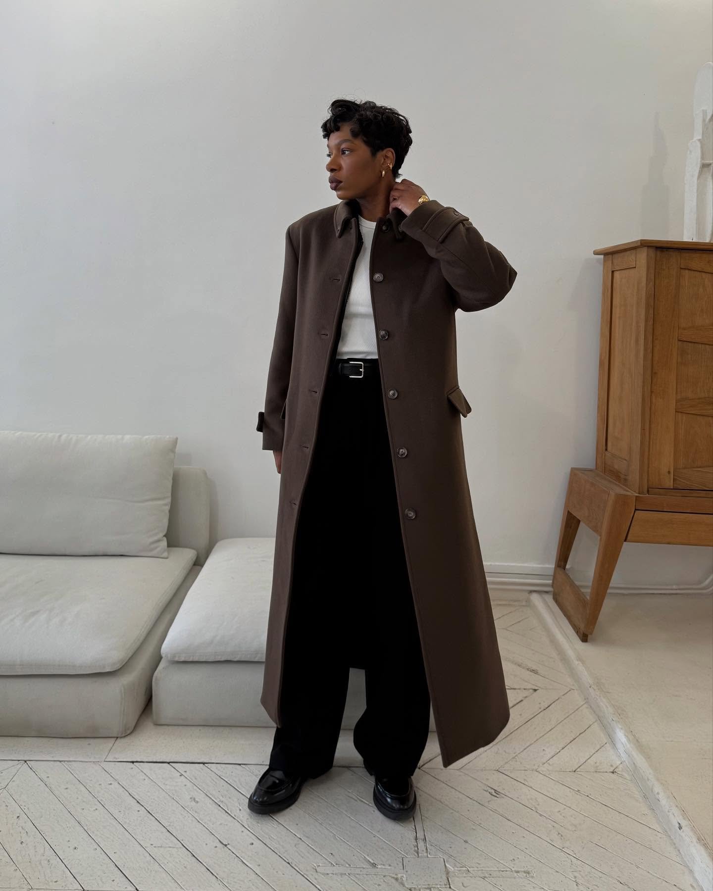 Pessoa vestindo sobretudo marrom elegante sobre roupa preta e blusa branca em sala minimalista com sofá.