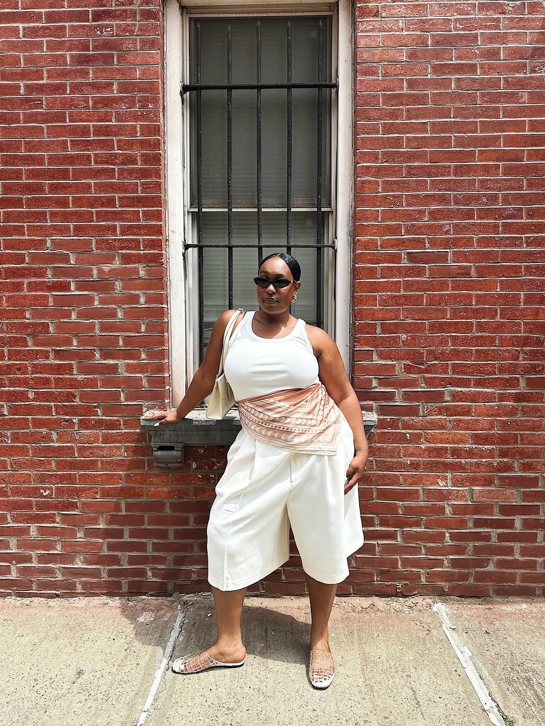 Pessoa com óculos de sol, roupa branca e sandálias, encostada em parede de tijolos. Looks de calor estilosos.