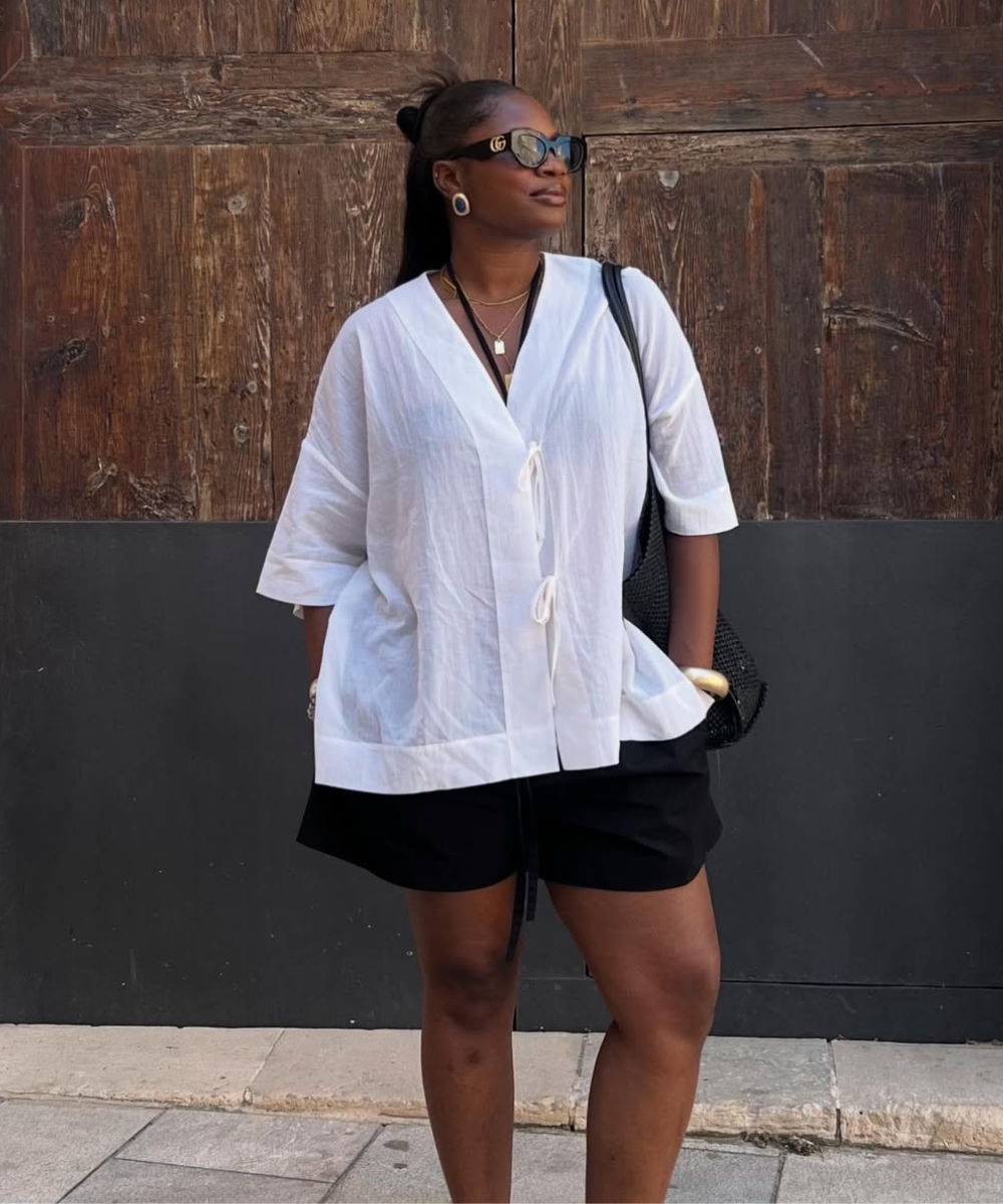 Pessoa com blusa branca e bermuda preta, usa óculos escuros, posando em frente a uma porta de madeira. Verão europeu 2026.