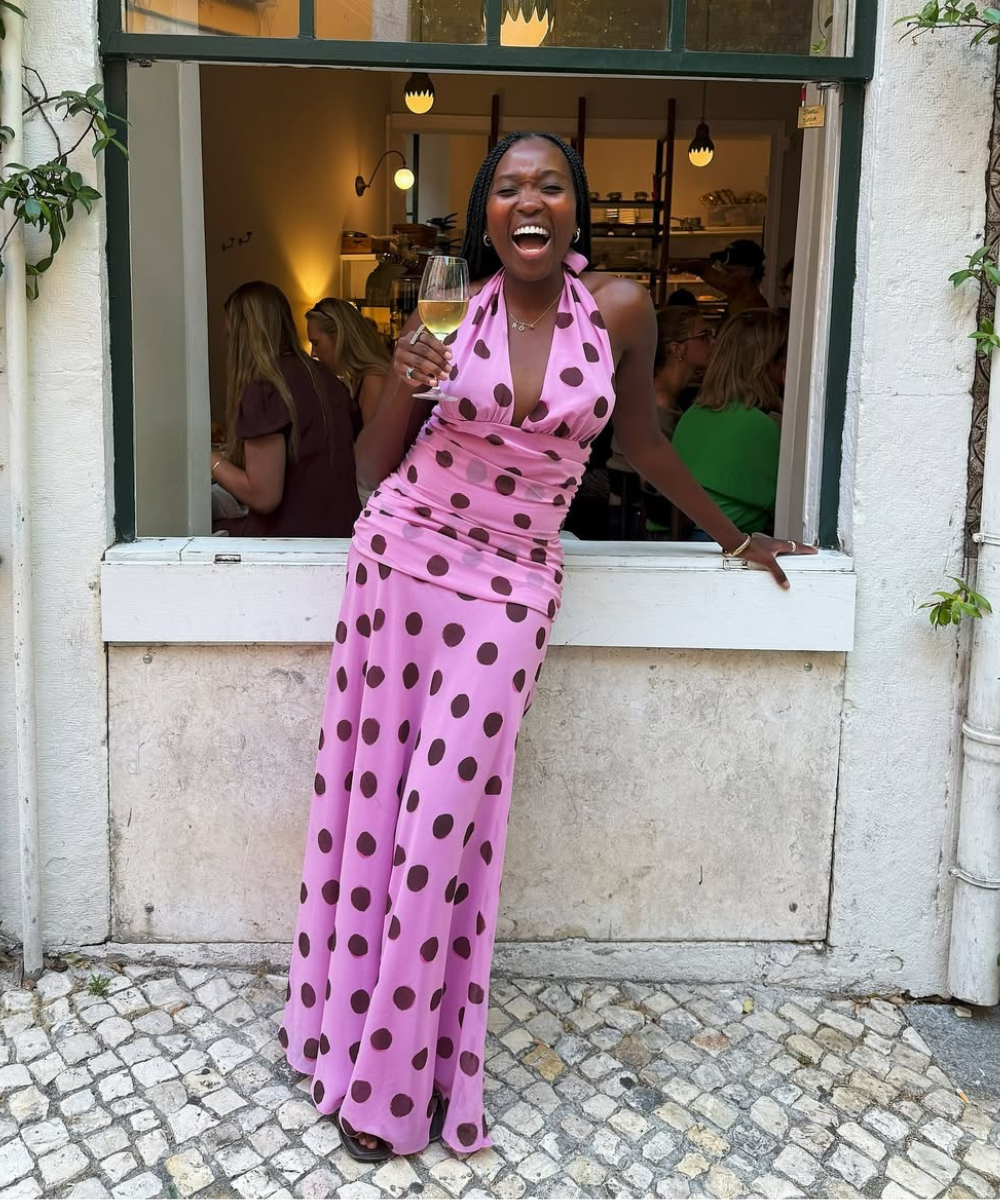 Mulher sorrindo em frente a uma janela, segurando uma taça de vinho, usando vestido rosa com bolinhas, verão europeu 2026.