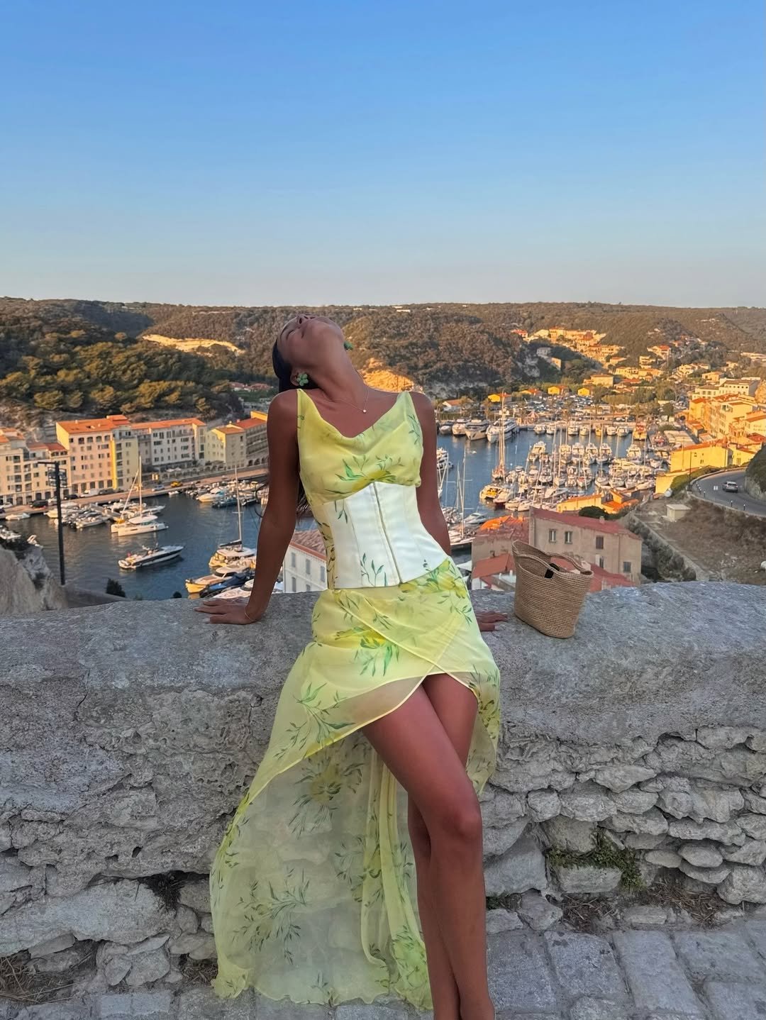 Mulher em vestido amarelo com corset branco, sentada em uma mureta com vista para uma marina ensolarada.