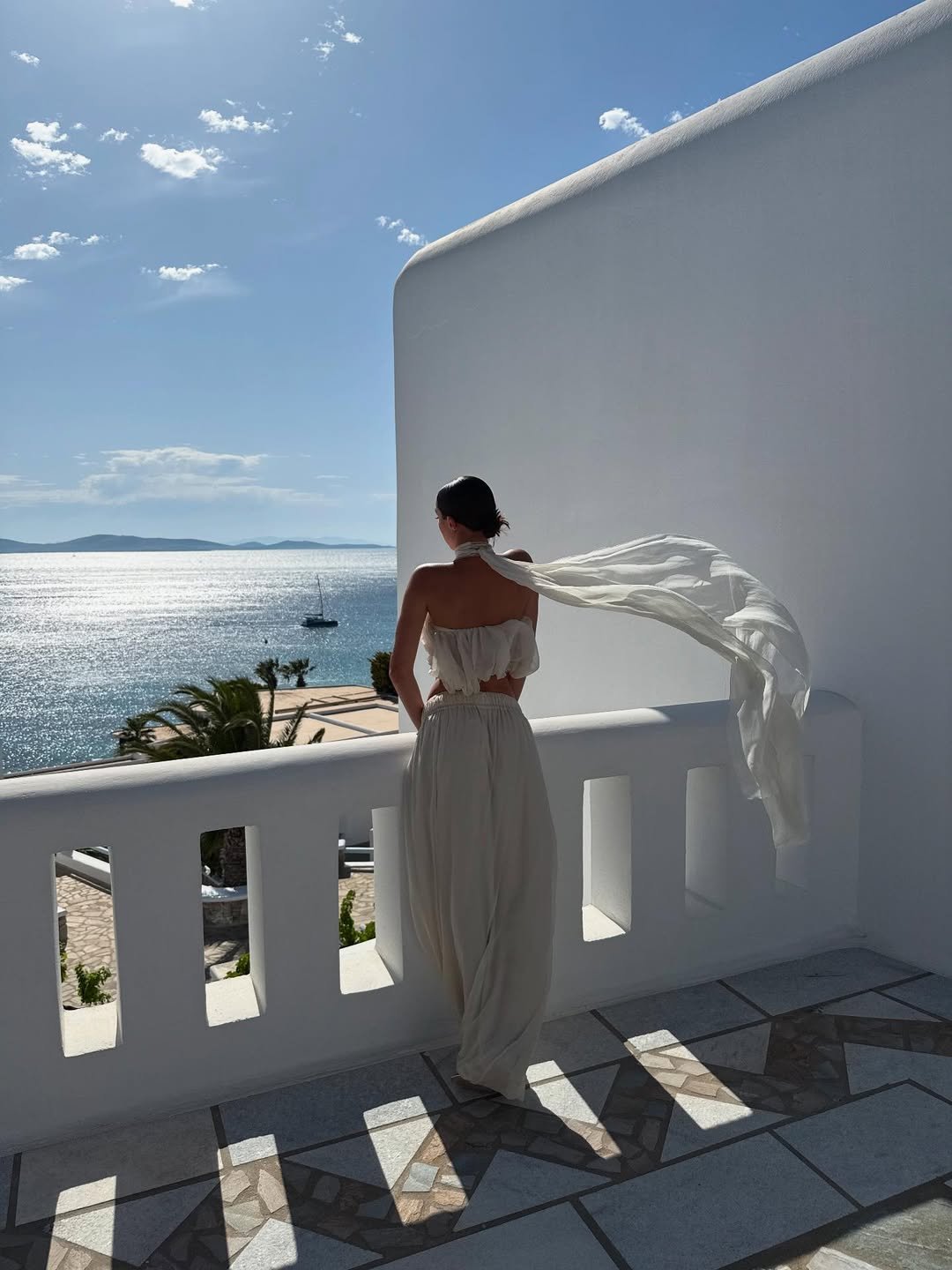 Pessoa de vestido fluido branco em uma varanda com vista para o mar. Ambiente evoca sofisticação e tranquilidade.