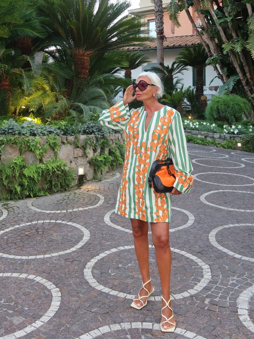 Mulher em vestido colorido posa ao ar livre com bolsa elegante, rodeada por palmeiras exuberantes. Estilo maximalista italiano.