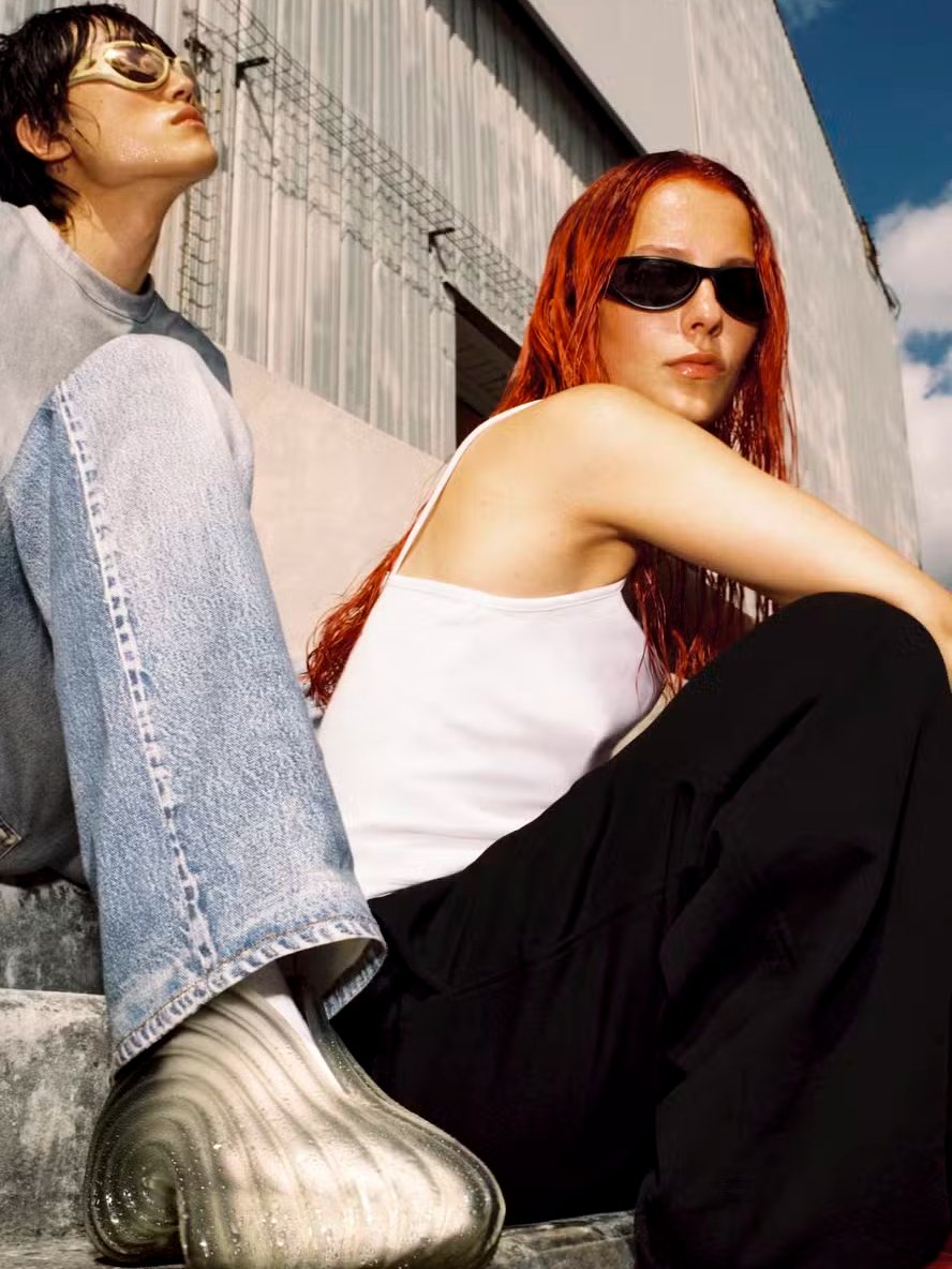 Há duas pessoas sentadas em uma escada. Uma delas veste óculos escuros. Sapatos Melissa aparecem.