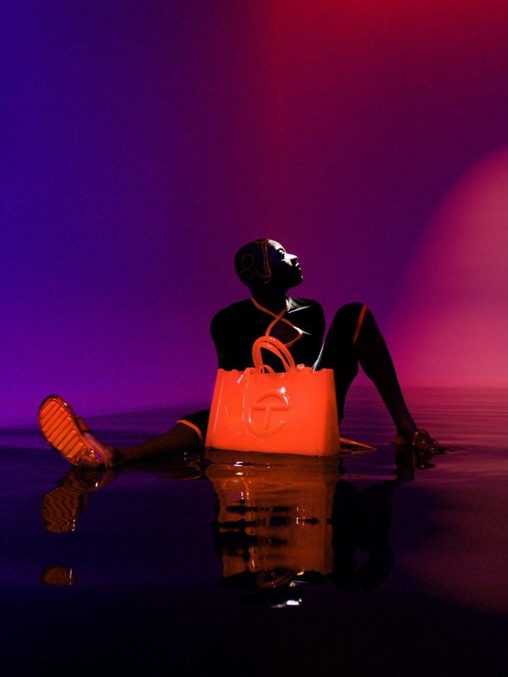 Modelo sentado na água, iluminado por luzes violeta e laranja, segurando uma bolsa laranja. Sandálias Melissa.
