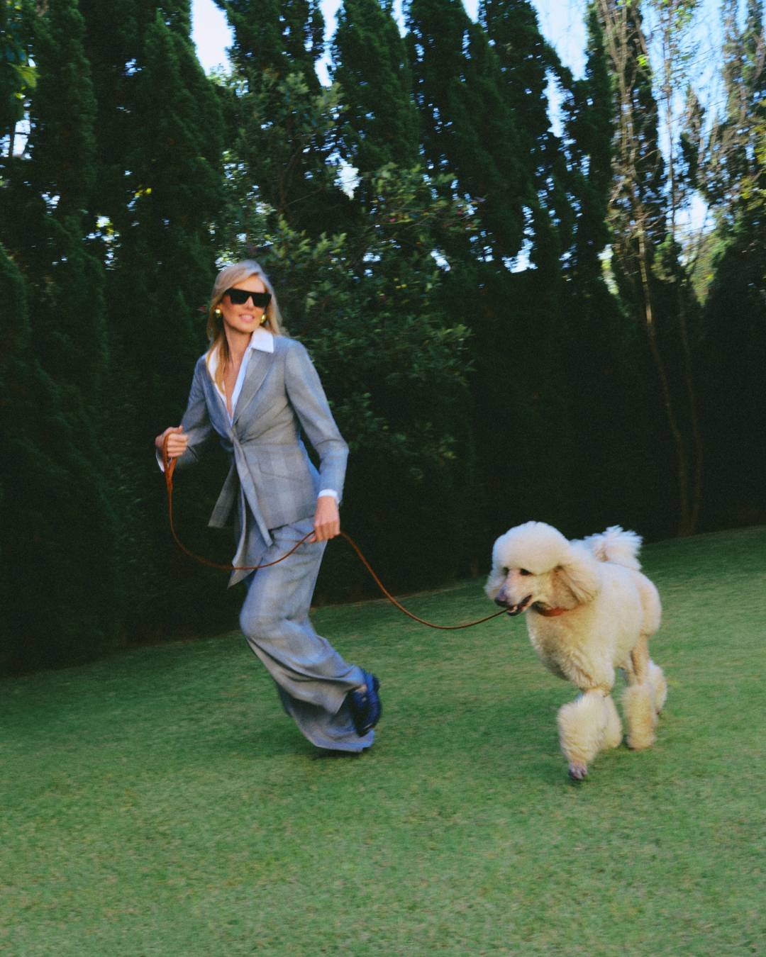 Pessoa em traje elegante, inspirado na moda dos anos 80 e 90, passeia com um cachorro em um jardim.