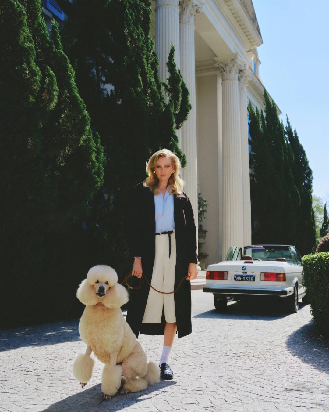 Pessoas em frente a uma mansão clássica com roupas elegantes e um poodle, evocando moda dos anos 80 e 90.