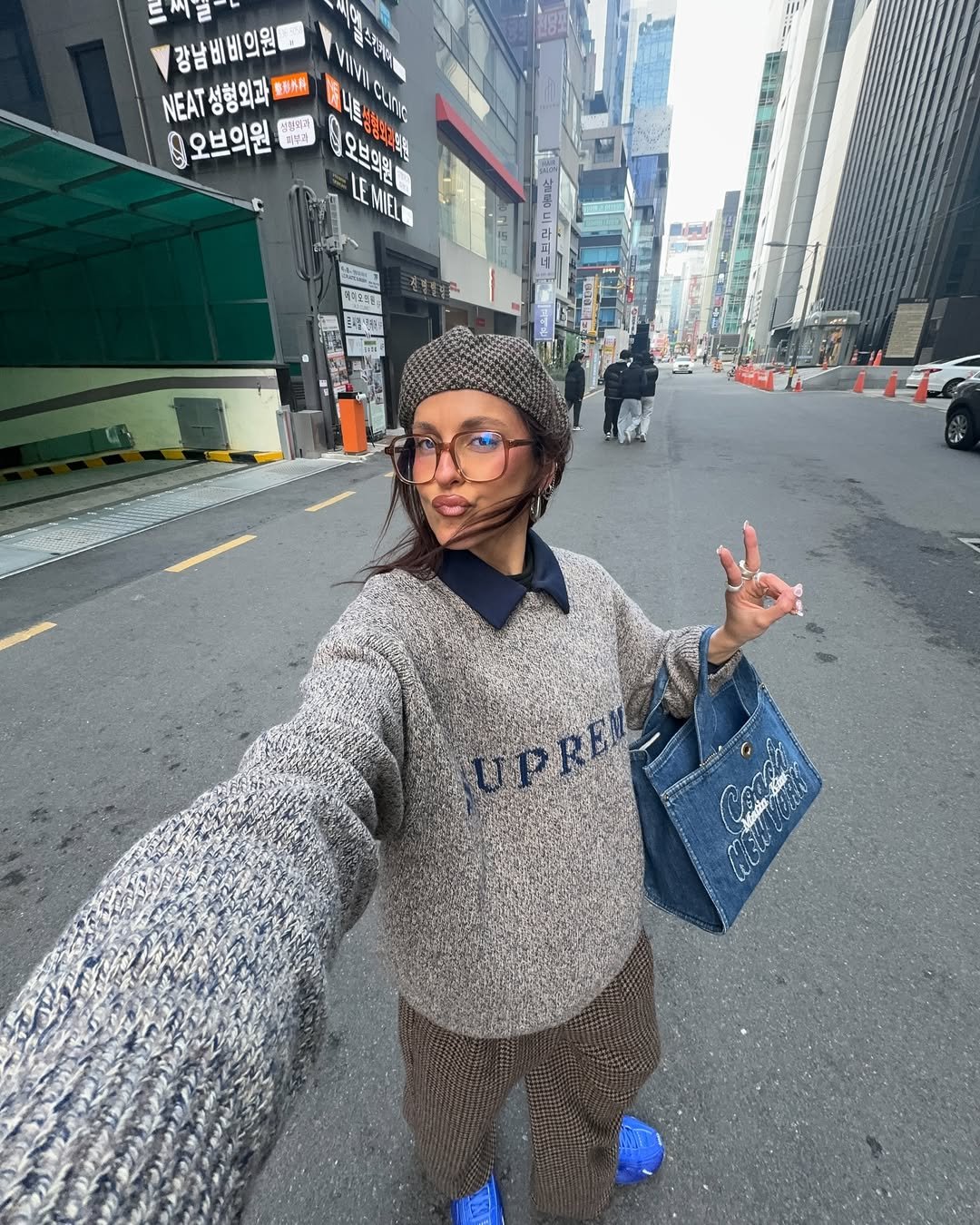 Mulher usando óculos de grau grandes em uma rua urbana, vestindo gorro e suéter, faz sinal de 