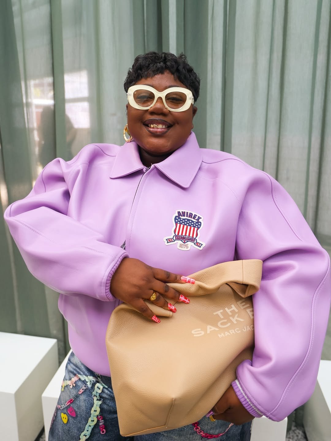 Pessoa sorridente com óculos de grau diferentes, usando casaco lilás com emblema, segura uma bolsa bege.