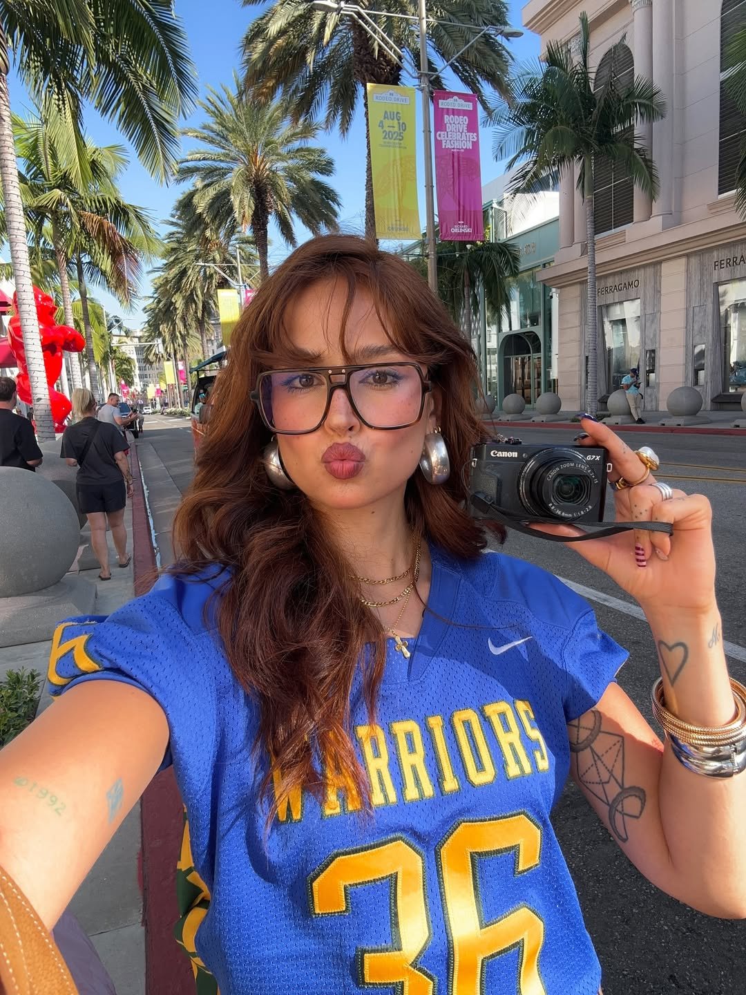 Pessoa usando óculos de grau grandes, camiseta azul, tirando selfie em rua arborizada com bandeiras coloridas ao fundo.