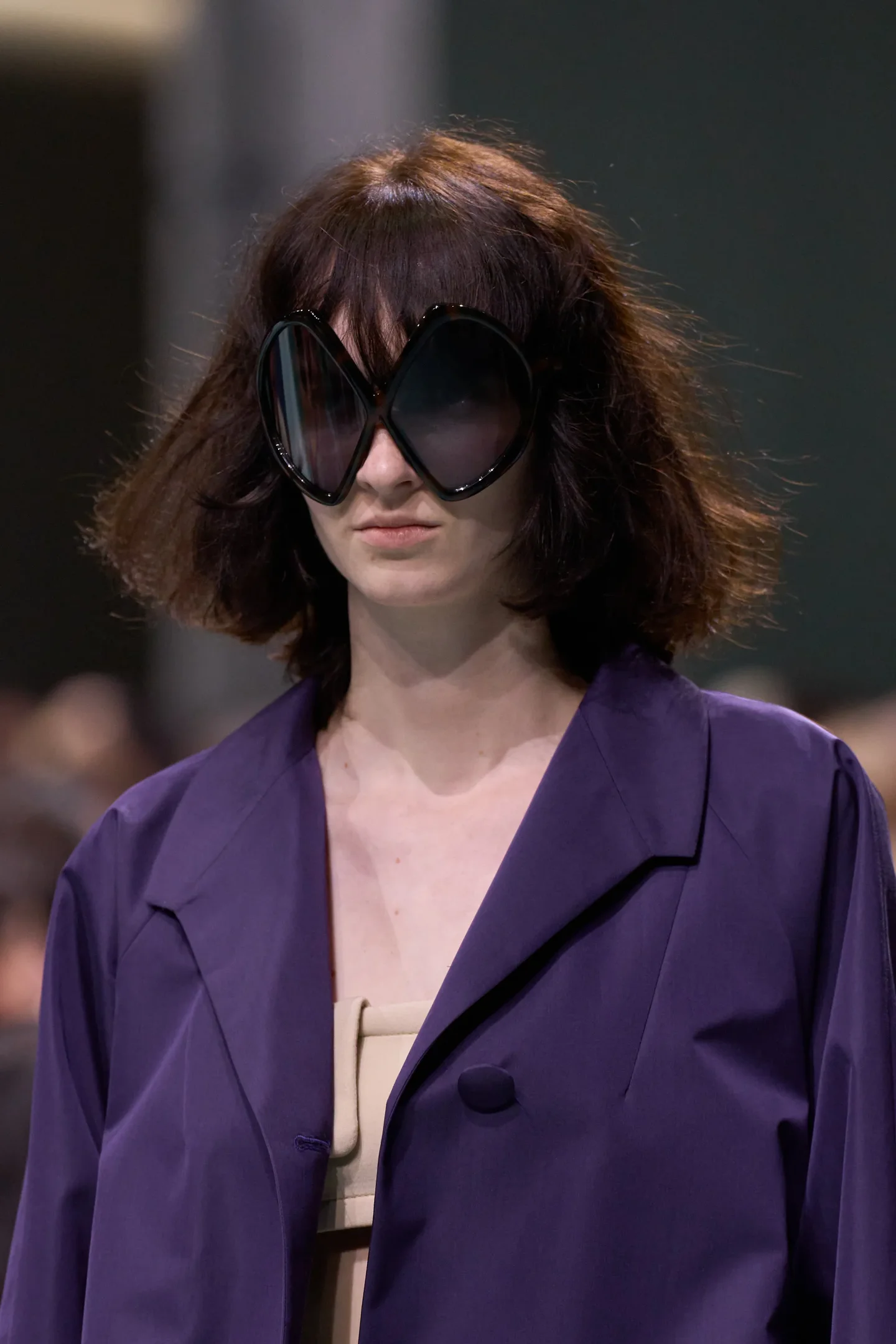 Modelo usando óculos Prada de sol grandes em formato inusitado, vestindo casaco roxo e vestido bege.