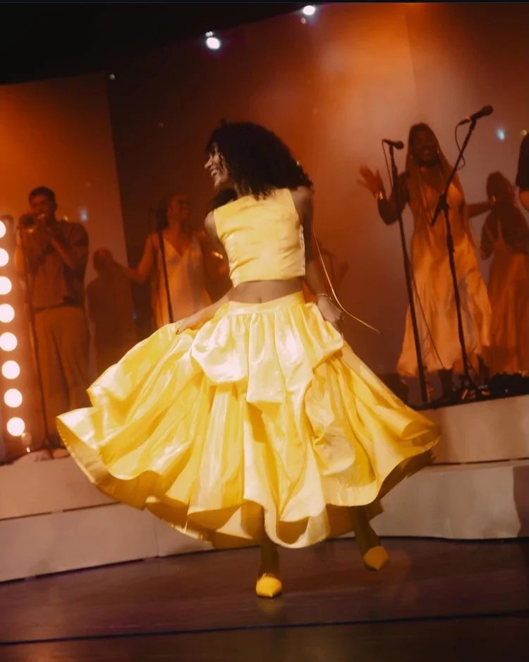 Olivia Dean dançando com vestido amarelo vibrante em um palco iluminado.