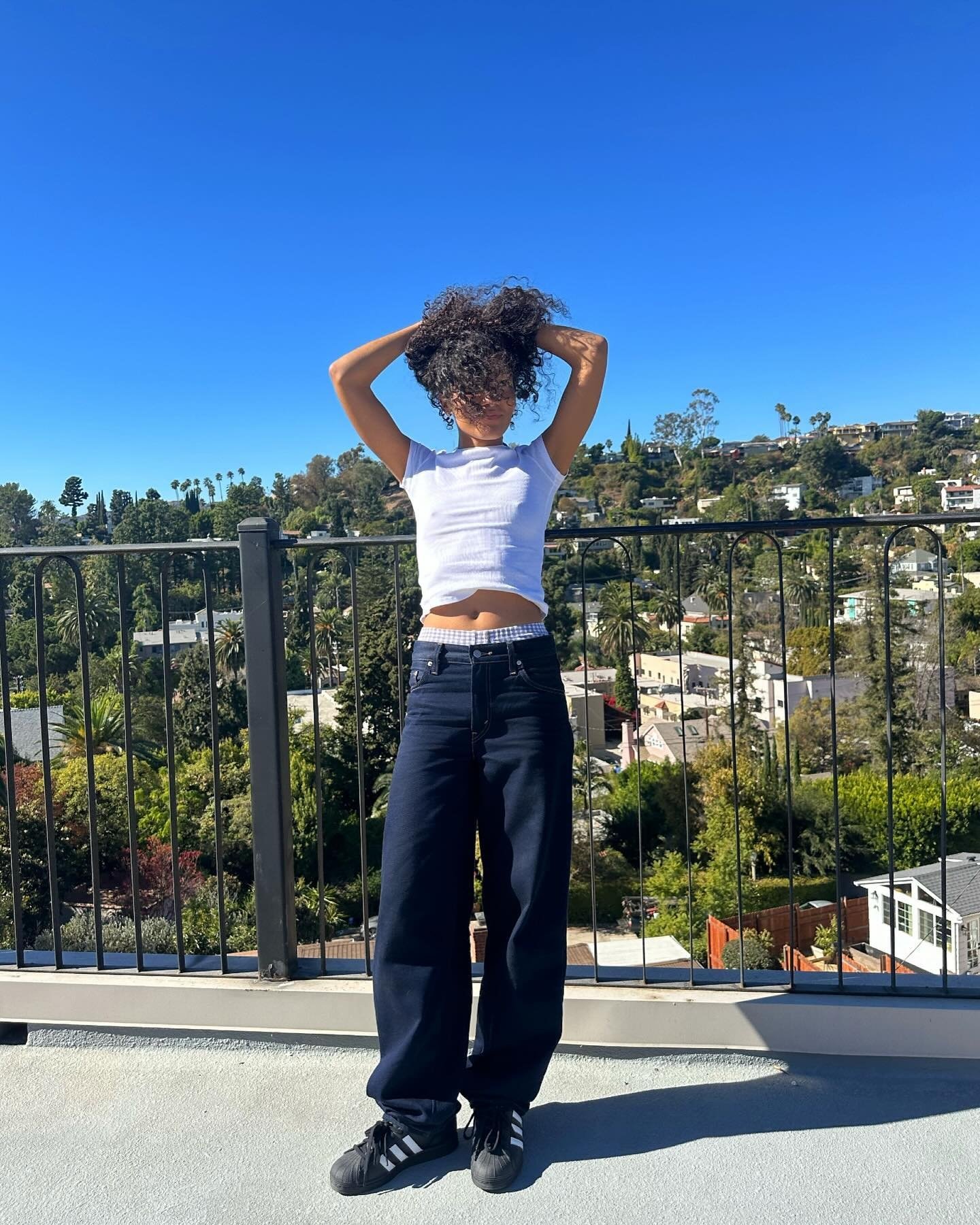 A imagem mostra uma mulher em uma varanda, sob um céu azul, com árvores ao fundo.
