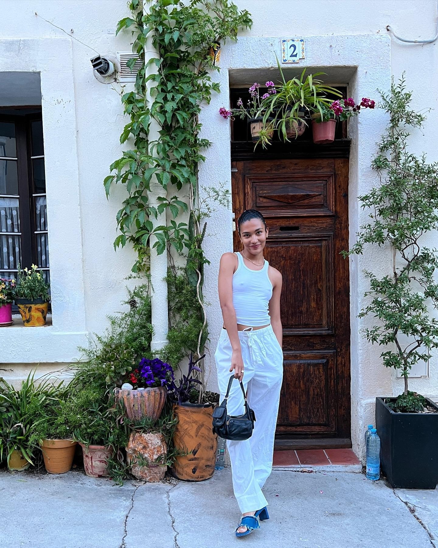 Pessoa de pé em frente a uma porta rústica, cercada por plantas diversas.