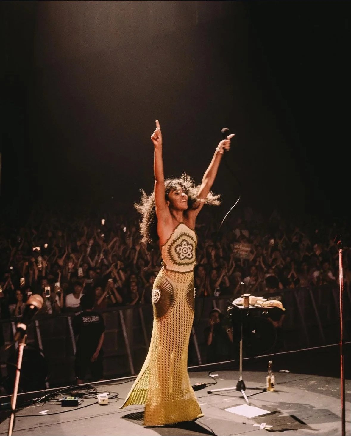 Cantora se apresenta animadamente em um palco diante de uma plateia cheia, com um vestido longo e microfone na mão.