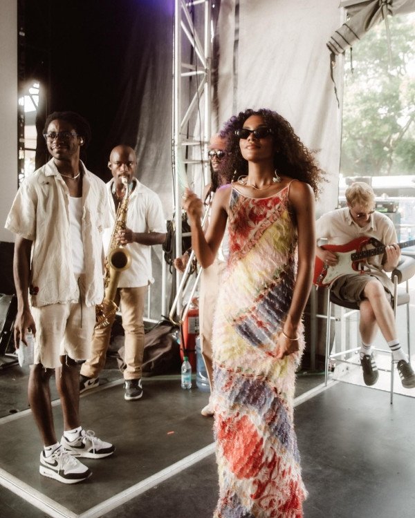 Grupo musical se prepara para apresentação em um palco. Olivia Dean com vestido colorido lidera.