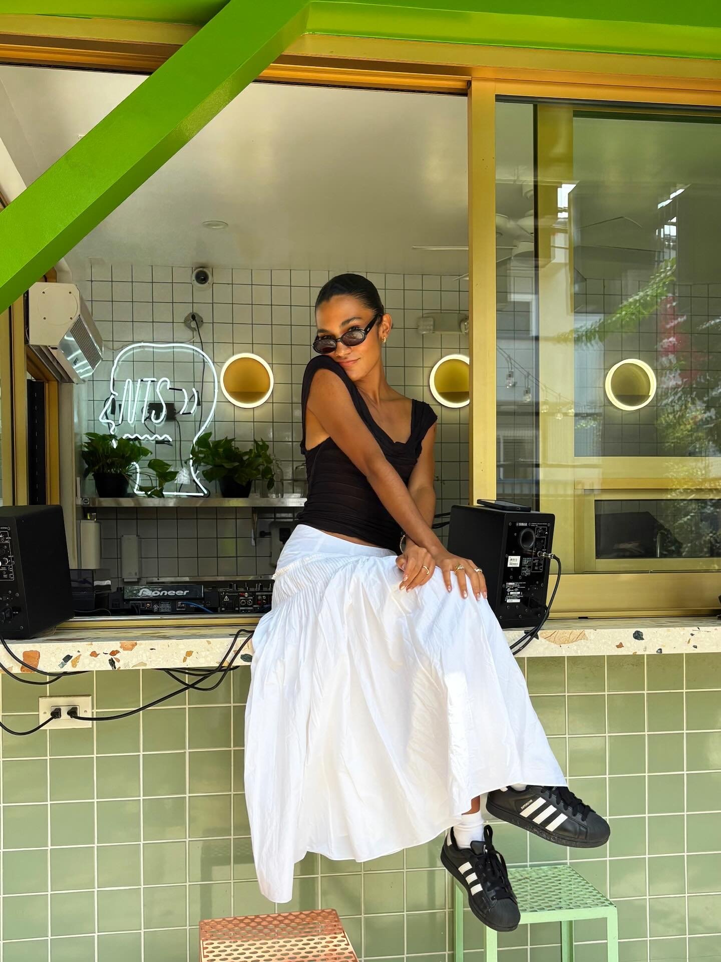 Pessoa com óculos escuros sentada em balcão verde, usando blusa preta, saia branca e tênis Adidas.