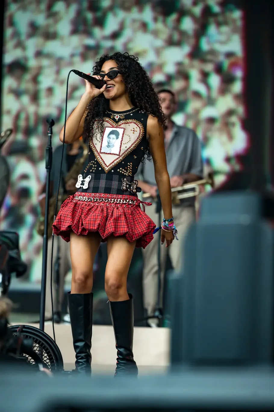 Cantora de óculos e saia xadrez canta em palco com fundo colorido.