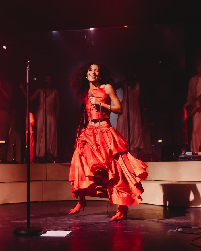 Cantora sorridente em um vestido vermelho vibrante performa no palco com microfone na mão.
